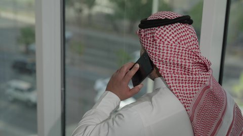 إجراء مكالمة هاتفية عبر الجوال، جهاز تقني حديث ومتطور،  تصوير مقرب من الخلف لرجل عربي خليجي سعودي يرتدي الثوب والشماغ التقليدي يقف داخل المبنى يقوم باستخدام الهاتف المحمول