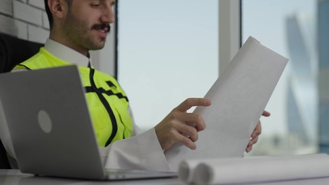 إدارة المشاريع الهندسية، متابعة استراتيجيات المشروع، مهن ووظائف سعودية، مفهوم الهندسة والإنشاءات المعمارية، مهندس عربي خليجي سعودي يرتدي سترة الحماية يفتح ويقراء المخطط الهندسي، خلفية أبراج وناطحات السحاب، تدقيق ومتابعة العمل