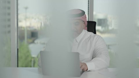 Gestures of integration and focus, providing a comfortable and suitable work environment, using a laptop to complete business tasks professionally, a Saudi Arabian Gulf man wearing a traditional thobe and shemagh sitting in an office working on a laptop, employment in Saudi offices and companies