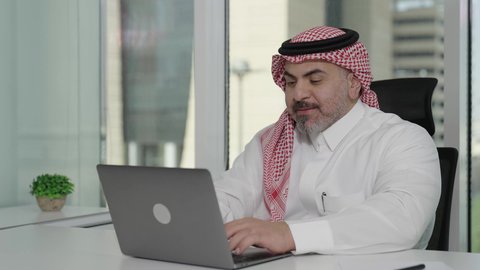 إيماءات الاندماج والتركيز، توفير بيئة عمل مريحة ومناسبة، استخدام جهاز الحاسوب المحمول لإنجاز مهام الاعمال باحترافية، رجل عربي خليجي سعودي يرتدي الثوب والشماغ التقليدي يجلس في المكتب يقوم بالعمل عبر اللابتوب، التوظيف في المكاتب والشركات السعودية