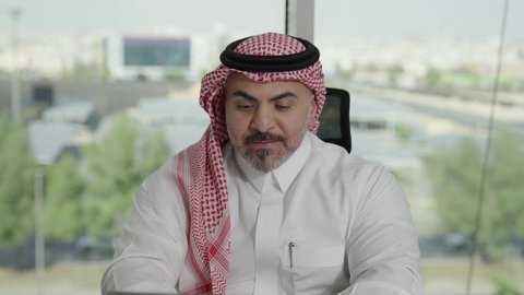 توفير بيئة عمل مريحة ومناسبة، استخدام جهاز الحاسوب المحمول لإنجاز مهام الاعمال باحترافية، رجل عربي خليجي سعودي يرتدي الثوب والشماغ التقليدي يجلس في المكتب يقوم بالعمل عبر اللابتوب، التوظيف في المكاتب والشركات السعودية