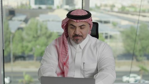 توفير بيئة عمل مريحة ومناسبة، استخدام جهاز الحاسوب المحمول لإنجاز مهام الاعمال باحترافية، رجل عربي خليجي سعودي يرتدي الثوب والشماغ التقليدي يجلس في المكتب يقوم بالعمل عبر اللابتوب، التوظيف في المكاتب والشركات السعودية