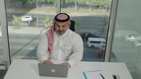 Office jobs and professions, gestures of integration and focus, providing a comfortable and suitable work environment, using a laptop to complete tasks professionally, a Saudi Arabian Gulf man wearing a traditional thobe and shemagh sitting in an office working on a laptop, employment in Saudi offices and companies