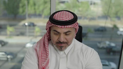 Office jobs and professions, gestures of integration and focus, providing a comfortable and suitable work environment, using a laptop to complete tasks professionally, a Saudi Arabian Gulf man wearing a traditional thobe and shemagh sitting in an office working on a laptop, employment in Saudi offices and companies