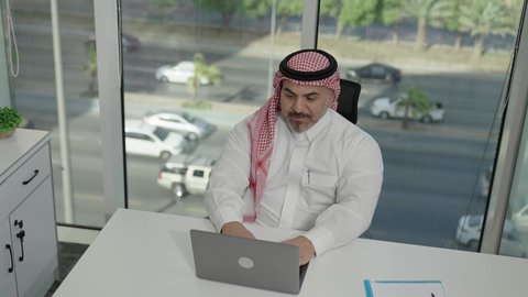 مهن وأعمال مكتبية، إيماءات الاندماج والتركيز، توفير بيئة عمل مريحة ومناسبة، استخدام جهاز الحاسوب المحمول لإنجاز المهام باحترافية، رجل عربي خليجي سعودي يرتدي الثوب والشماغ التقليدي يجلس في المكتب يقوم بالعمل عبر اللابتوب، التوظيف في المكاتب والشركات السعودية
