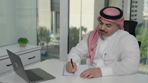 Office jobs and professions, gestures of integration and focus, providing a comfortable and suitable work environment, using a laptop to complete tasks professionally, a Saudi Arabian Gulf man wearing a traditional thobe and shemagh sitting in an office taking notes, employment in Saudi offices and companies