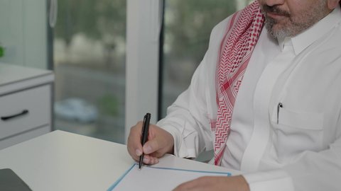 مهن وأعمال مكتبية، إيماءات الاندماج والتركيز، توفير بيئة عمل مريحة ومناسبة، استخدام جهاز الحاسوب المحمول لإنجاز المهام باحترافية، رجل عربي خليجي سعودي يرتدي الثوب والشماغ التقليدي يجلس في المكتب يقوم بتدوين الملاحظات، التوظيف في المكاتب والشركات السعودية