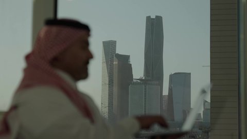 إيماءات الاندماج والتركيز، استخدام جهاز الحاسوب المحمول لإنجاز المهام باحترافية، رجل عربي خليجي سعودي يرتدي الثوب والشماغ التقليدي يحمل اللابتوب، التوظيف في المكاتب والشركات السعودية، خلفية الأبراج وناطحات السحاب في مدينة الرياض