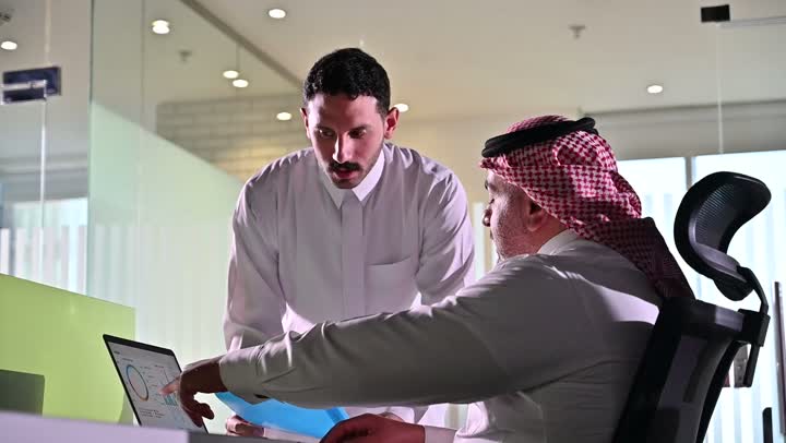 استخدام الحاسب والتقنية في مجال الاعمال، بيئة العمل المريحة في الشركات السعودية، الحوار و النقاش بين الزملاء، تبادل الخبرات والمعلومات لتحقيق الاهداف, موظفان عربيان خليجيان سعوديان يجلسان على مكتبهم داخل مقر الشركة