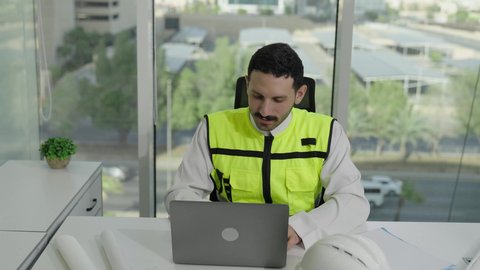 تطور السعودية على يد مهندسين الوطن، مهن ووظائف سعودية، مفهوم الهندسة والإنشاءات المعمارية، مهندس عربي خليجي سعودي يرتدي سترة الحماية ينجز المهام اليومية على مكتب العمل، متابعة سير المشروع من خلال الحاسوب المحمول، دمج العمل مع التكنولولجيا