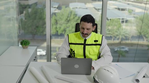 تطور السعودية على يد مهندسين الوطن، مهن ووظائف سعودية، مفهوم الهندسة والإنشاءات المعمارية، مهندس عربي خليجي سعودي يرتدي سترة الحماية ينجز المهام اليومية على مكتب العمل، متابعة سير المشروع من خلال الحاسوب المحمول، دمج العمل مع التكنولولجيا
