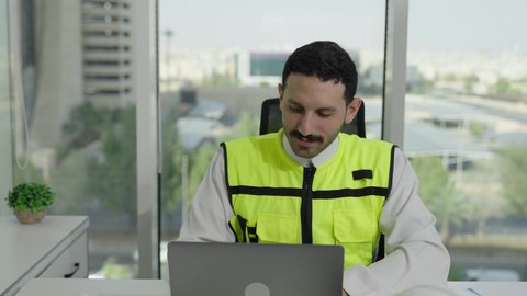  مفهوم الهندسة والإنشاءات المعمارية، تطور السعودية على يد مهندسين الوطن، مهن ووظائف سعودية، تصوير مقرب لمهندس عربي خليجي سعودي يرتدي سترة الحماية ينجز المهام اليومية على مكتب العمل، متابعة سير المشروع من خلال الحاسوب المحمول، دمج العمل مع التكنولولجيا