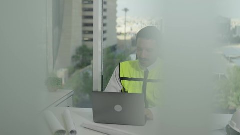 The concept of engineering and architectural construction, the development of Saudi Arabia at the hands of the nation's engineers, Saudi professions and jobs, a Saudi Arabian Gulf engineer wearing a protective vest performing daily tasks at the work desk, monitoring the progress of the project via a laptop, integrating work with technology.
