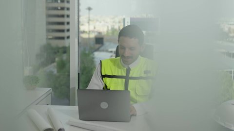 مفهوم الهندسة والإنشاءات المعمارية، تطور السعودية على يد مهندسين الوطن، مهن ووظائف سعودية،مهندس عربي خليجي سعودي يرتدي سترة الحماية ينجز المهام اليومية على مكتب العمل، متابعة سير المشروع من خلال الحاسوب المحمول، دمج العمل مع التكنولولجيا