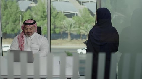 استخدام جهاز تقني حديث، موظفة تقوم بتقديم المشاريع للمدير لتدقيقها عبر الجهاز اللوحي، تطوير ودراسة وتحليل المشاريع الجديدة، موظف عربي خليجي سعودي  يرتدي الثوب والشماغ التقليدي يجلس خلف المكتب داخل مقر الشركة، الوحدة والعمل الجماعي، مهنة ووظيفة سعودية مكتبية