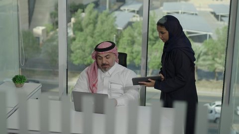 استخدام جهاز تقني حديث، موظفة تقوم بتقديم المشاريع للمدير لتدقيقها عبر الجهاز اللوحي، تطوير ودراسة وتحليل المشاريع الجديدة، موظف عربي خليجي سعودي  يرتدي الثوب والشماغ التقليدي يجلس خلف المكتب داخل مقر الشركة، الوحدة والعمل الجماعي، مهنة ووظيفة سعودية مكتبية