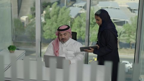 استخدام جهاز تقني حديث، موظفة تقوم بتقديم المشاريع للمدير لتدقيقها عبر الجهاز اللوحي، تطوير ودراسة وتحليل المشاريع الجديدة، موظف عربي خليجي سعودي  يرتدي الثوب والشماغ التقليدي يجلس خلف المكتب داخل مقر الشركة، الوحدة والعمل الجماعي، مهنة ووظيفة سعودية مكتبية