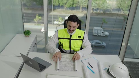 الخطط والمشاريع الهندسية، تطور السعودية على يد مهندسين الوطن، مهن ووظائف سعودية، تصوير علوي لمهندس عربي خليجي سعودي يرتدي سترة الحماية يحمل ويقراء المخطط الهندسي، التدقيق والإشرف على العمل، مهندس معماري محترف، دمج العمل مع التكنولولجيا