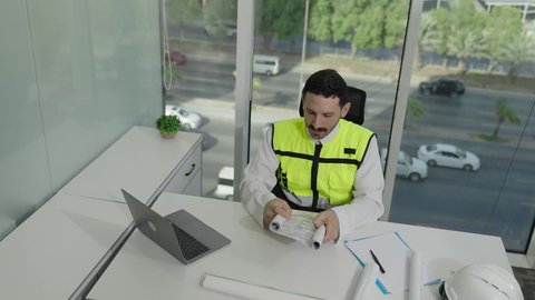 الخطط والمشاريع الهندسية، تطور السعودية على يد مهندسين الوطن، مهن ووظائف سعودية، تصوير علوي لمهندس عربي خليجي سعودي يرتدي سترة الحماية يحمل ويقراء المخطط الهندسي، التدقيق والإشرف على العمل، مهندس معماري محترف، دمج العمل مع التكنولولجيا