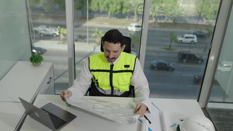 الخطط والمشاريع الهندسية، تطور السعودية على يد مهندسين الوطن، مهن ووظائف سعودية، تصوير علوي لمهندس عربي خليجي سعودي يرتدي سترة الحماية يحمل ويقراء المخطط الهندسي، التدقيق والإشرف على العمل، مهندس معماري محترف، دمج العمل مع التكنولولجيا