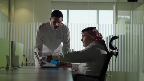 الوحدة والعمل الجماعي، استخدام جهاز تقني حديث، موظف يقوم بتقديم المشاريع للمدير لتدقيقها، تطوير ودراسة وتحليل المشاريع الجديدة، موظف عربي خليجي سعودي  يرتدي الثوب والشماغ التقليدي يجلس خلف المكتب داخل مقر الشركة، مهنة ووظيفة سعودية مكتبية
