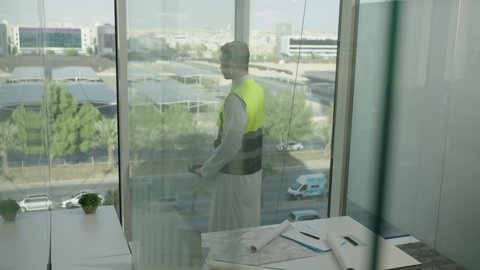 تصويرجانبي بطيئ لمهندس عربي خليجي سعودي يرتدي سترة الحماية والخوذة البيضاء يتأمل معالم المدينة، مفهوم الهندسة المعمارية، المهن والوظائف الشبابية السعودية، تطور ونمو القطاع الهندسي، خلفية حركة المرور