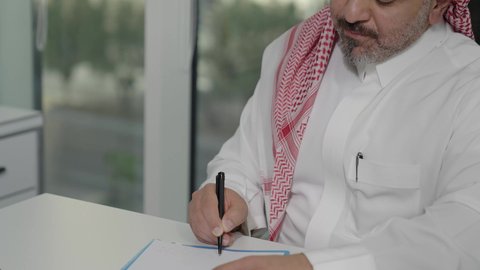 مفهوم ادارة الاعمال، كتابة وتدوين الملاحظات المهمة، مهن ووظائف مكتبيه في الممكلة العربية السعودية, تصوير مقرب لرجل عربي خليجي سعودي يرتدي الثوب والشماغ التقليدي يجلس خلف طاولة المكتب بإيماءات الاندماج والتركيز