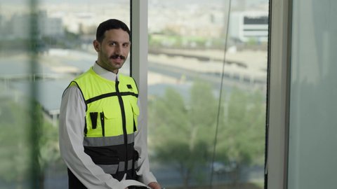 المهن والوظائف الشبابية السعودية، تصويرمقرب لمهندس عربي خليجي سعودي مبتسم يرتدي خوذة بيضاء وسترة الحماية ، مفهوم الهندسة المعمارية،  تطور ونمو القطاع الهندسي، خلفية حركة المرور ومعالم المدينة، الوقوف بثقة والنظر إلى الكاميرا