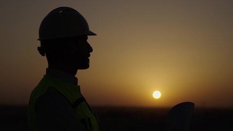 الإشراف على العمل وتدقيقه، المهن والوظائف الشبابية السعودية، تصويرجانبي مقرب لمهندس عربي خليجي سعودي يرتدي خوذة وسترة الحماية يحمل ويقراء المخطط الهندسي، مفهوم الهندسة المعمارية، تطور ونمو القطاع الهندسي، خلفية غروب الشمس ومعالم المدينة، الوقوف بثقة وثبات