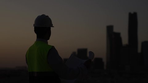الإشراف على العمل وتدقيقه، المهن والوظائف الشبابية السعودية، تصويرخلفي لمهندس عربي خليجي سعودي يرتدي خوذة وسترة الحماية يحمل ويقراء المخطط الهندسي، مفهوم الهندسة المعمارية، تطور ونمو القطاع الهندسي، خلفية غروب الشمس ومعالم المدينة، الوقوف بثقة وثبات