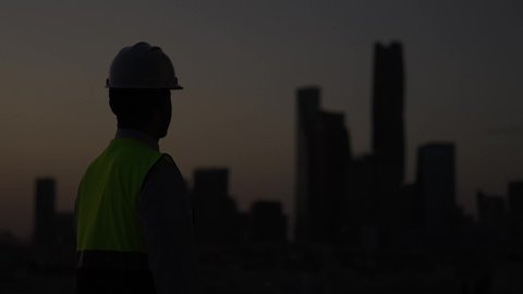 الإنشاءات والهندسة المعمارية، المهن والوظائف الشبابية السعودية، تصويرخلفي بطيئ لمهندس عربي خليجي سعودي يرتدي خوذة وسترة الحماية يتأمل معالم المدينة، تطور ونمو القطاع الهندسي، خلفية غروب الشمس، الوقوف بثقة وثبات