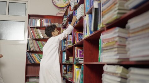قضاء وقت ممتع ، ممارسة الهوايات المفضلة ، طالب عربي سعودي يقف بجوار مكتبة خشبية في المكتبة المدرسية، الاستمتاع بقراءة الكتب المدرسية، التعليم في المملكة العربية السعودية