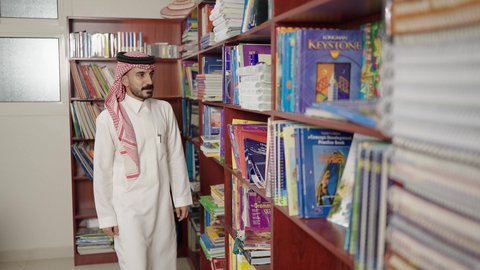 قضاء وقت ممتع ، ممارسة الهوايات المفضلة ، معلم عربي سعودي يقف بجوار مكتبة خشبية في المكتبة المدرسية، الاستمتاع بقراءة الكتب المدرسية، التعليم في المملكة العربية السعودية
