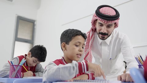 Education and teaching with the prescribed curricula for students, a Gulf Arab Saudi teacher wearing traditional attire and a shemagh discussing lessons with his students, active participation of students with teachers, the quality of education in Saudi Arabia, friendly dialogues between the teacher and students.