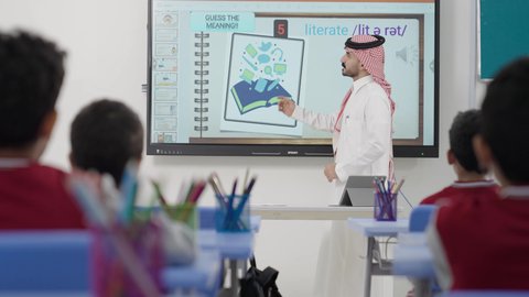 The development of teaching methods in the schools of the Kingdom, benefiting from advanced technologies in learning and studying, developed curricula, and e-learning. Two Saudi Gulf Arab students are sitting in their seats, and in front of them is a Saudi teacher using the blackboard to explain.