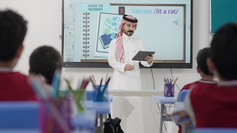 الاستفادة من التقنيات والتكنولوجيا الحديثة في التعلم والدراسة، المناهج المطورة والتعليم الالكتروني، تطور طرائق التدريس في مدارس المملكة، طلاب عربيين خليجيين سعوديين يجلسون في مقاعدهم وأمامهم مدرس سعودي يستخدم جهاز التابلت