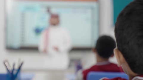 طالب عربي خليجي سعودي مبتسم يرتدي الثوب التقليدي يرفع يديه للإجابة، التفاعل والمشاركة داخل الفصل الدراسي، التربية والتعليم بالمناهج المقررة للطلاب في المملكة العربية السعودية، تعابير وايماءات الحماس لمعرفة الجواب الصحيح