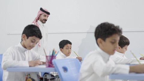 Friendly dialogues between the teacher and students, active participation of students with teachers, a Saudi Gulf Arab teacher wearing traditional attire and a shemagh discussing lessons with his students, the quality of education in Saudi Arabia, education and teaching with the prescribed curricula for students.