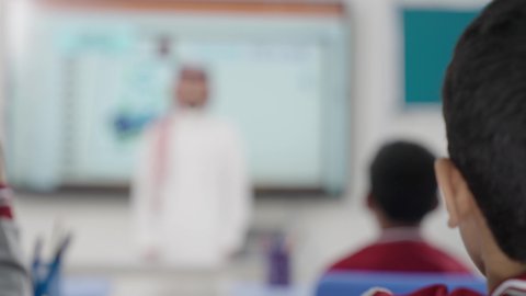 التفاعل والمشاركة داخل الفصل الدراسي، طالب عربي خليجي سعودي مبتسم يرتدي الثوب التقليدي يرفع يديه للإجابة، التربية والتعليم بالمناهج المقررة للطلاب في المملكة العربية السعودية، تعابير وايماءات الحماس لمعرفة الجواب الصحيح
