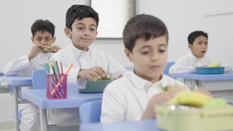 الاستمتاع بتناول الأطعمة الصحية في وقت الفسحة المدرسية، موسم العودة للمدارس، مجموعة من الطلاب العربيين الخليجيين السعوديين   يجلسون في مقاعدهم وأمامهم اللانش البوكس، الوجبات الغنية بالعناصر الغذائية الهامة لبناء الجسم
