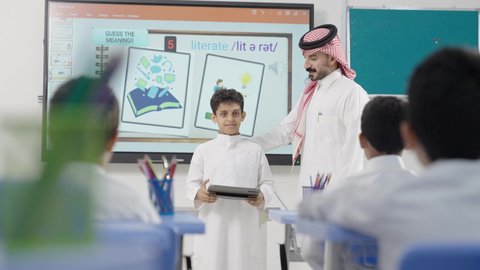 المناهج المطورة والتعليم الالكتروني، الاستفادة من التقنيات والتكنولوجيا الحديثة في التعلم والدراسة، تطور طرائق التدريس في مدارس المملكة، طلاب عربيين خليجيين سعوديين  يجلسون في مقاعدهم وأمامهم طالب يستخدم جهاز التابلت