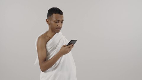 تطبيق مرشد الحج الالكتروني، استخدام خدمة تطبيق الارشادات لأداء مناسك الحج والعمرة، بورتريه لرجل مسلم خليجي عربي سعودي يرتدي ملابس الإحرام يستخدم الهاتف المحمول، مفهوم أركان الإسلام، خلفية رمادي