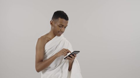 Using the Hajj and Umrah Guide application service, portrait of a smiling Saudi Arabian Gulf Muslim man wearing Ihram clothes using a mobile phone, electronic Hajj Guide application, concept of the pillars of Islam, gray background