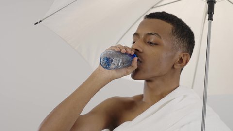 الركن الخامس من أركان الاسلام، ايماءات البهجة والارتياح، زيارة بيت الله الحرام في مكة المكرمة، بورتريه لرجل عربي خليجي سعودي يرتدي ملابس الاحرام يحمل بيده مظلة للحماية من أشعة الشمس و يقوم بشرب الماء، آداء مناسك الحج والعمرة، خلفية رمادي