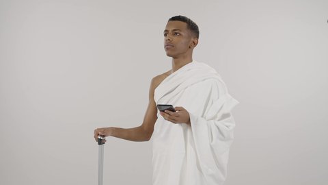 Knowing and determining the location via mobile phone, visiting the Holy House of God in Mecca, portrait of a Saudi Arabian Gulf man wearing Ihram clothes holding an umbrella and looking at a mobile phone, performing Hajj and Umrah rituals, gray background