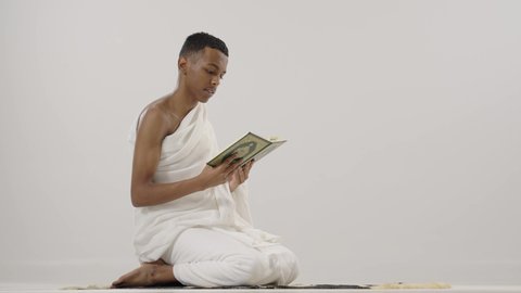 إيماءات الخشوع و الاجتهاد في العباده, قراءة القرآن الكريم، بورتريه لرجل عربي خليجي سعودي يرتدي لباس الإحرام يحمل بيده المصحف الشريف و يقوم بالتلاوة منه, الإجتهاد في العبادة, التقرب الى الله تعالى, مفهوم أركان الإسلام، خلفية رمادي