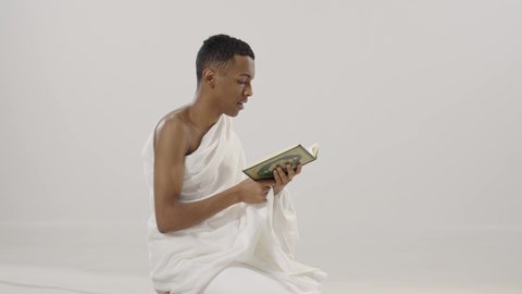 الحرص على عدم تفويت وقت الصلاة، التقرب إلى الله بالعبادة والطاعة، قراءة القرآن الكريم، بورتريه لرجل عربي خليجي سعودي يرتدي لباس الإحرام يحمل بيده المصحف الشريف و يقوم بالتلاوة منه, خلفية رمادي