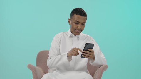إرسال الرسائل النصية والصوتية عبر الجوال، مفهوم الجلوس والراحة، استخدام الأجهزة والوسائل التقنية الحديثة، بورتريه لرجل عربي خليجي سعودي يرتدي الثوب التقليدي يجلس على كرسي مريح يمسك الهاتف المحمول بتعابير السعادة, خلفية زرقاء