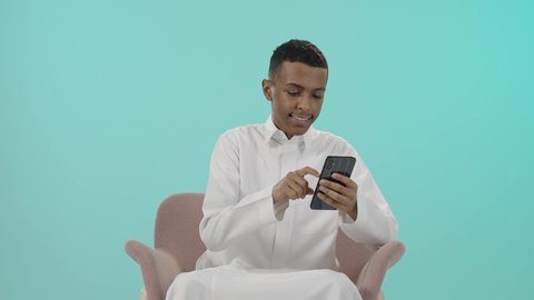 مفهوم الجلوس والراحة،  استخدام الأجهزة والوسائل التقنية الحديثة، إرسال الرسائل النصية والصوتية عبر الجوال، بورتريه لرجل عربي خليجي سعودي يرتدي الثوب التقليدي يجلس على كرسي مريح يمسك الهاتف المحمول بتعابير السعادة, خلفية زرقاء