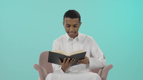 A calm atmosphere for concentration, the concept of diligence and excellence, preparing for the new school year, preparing lessons to achieve success, a portrait of a young Saudi Arabian Gulf man wearing traditional clothing reading a book, developing educational curricula in Saudi Arabia, blue background